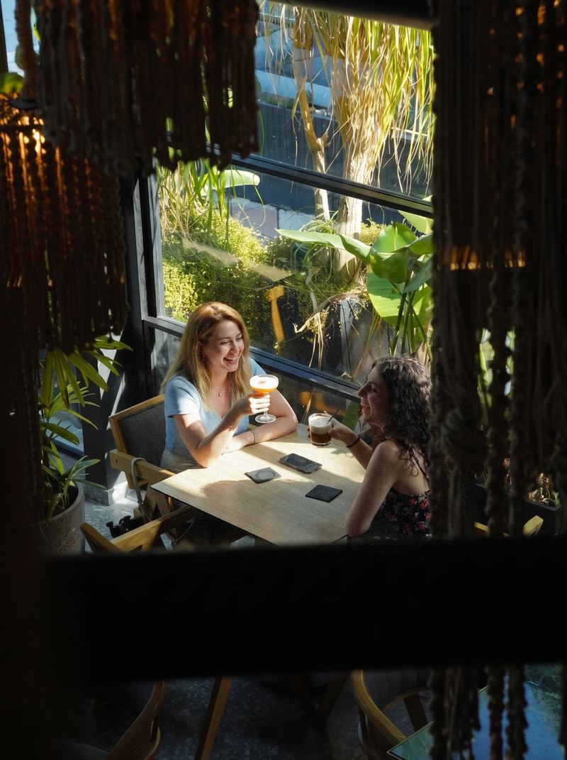Two guests enjoying cocktails by the window inside Al Hayat, framed by hanging macramé and lush greenery