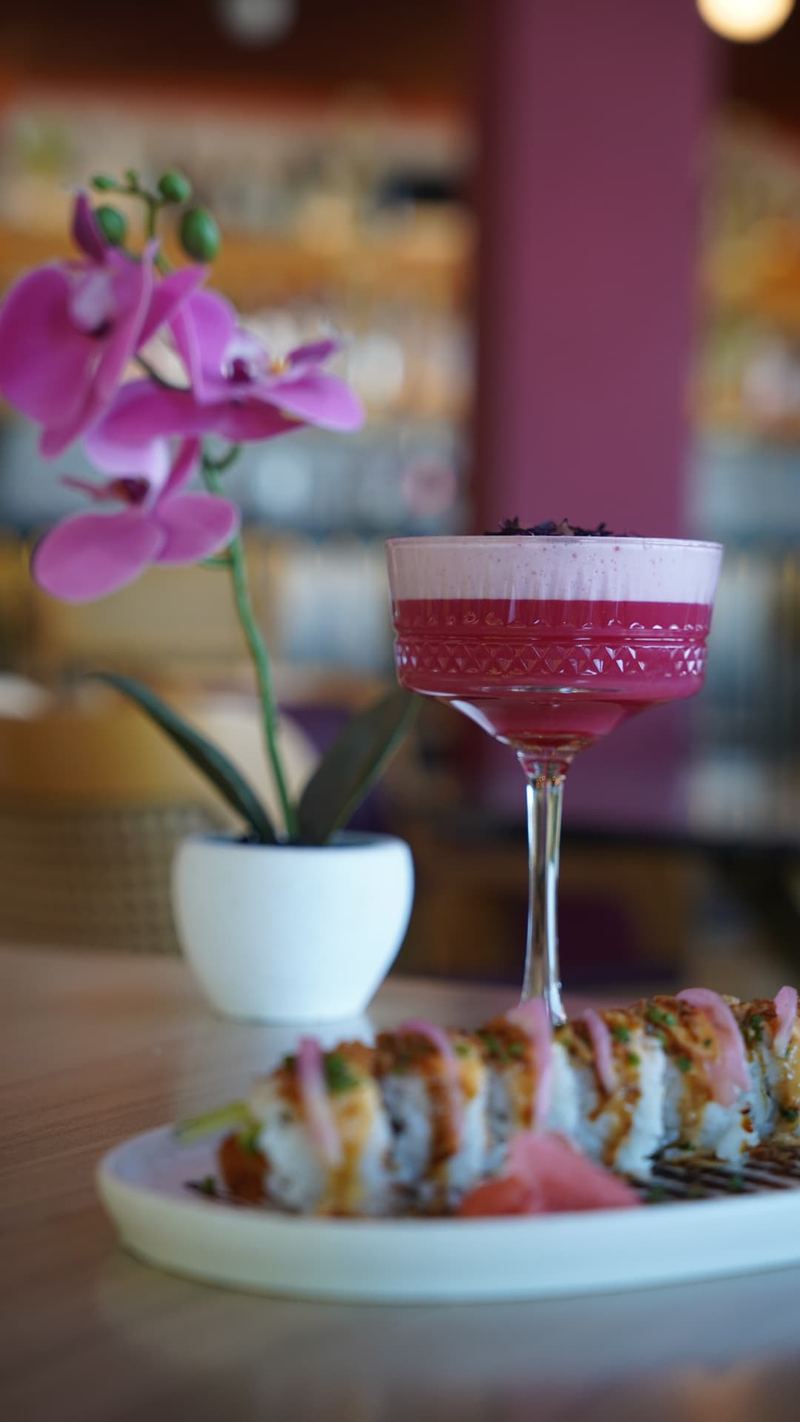 Tall crystal coupe holding a vivid pink cocktail beside a sushi roll platter and a potted orchid at the Al Hayat bar