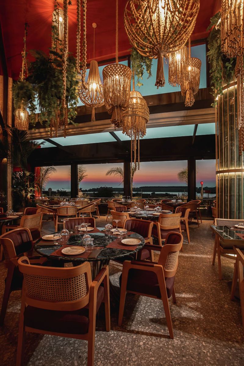 Dining room framed by floor-to-ceiling windows revealing a pink sunset over the Rhodes seafront and marina