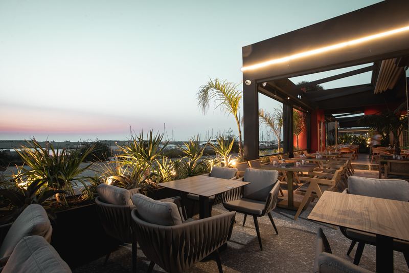 Al Hayat's outdoor terrace with lounge armchairs, illuminated planters and a marina sunset backdrop