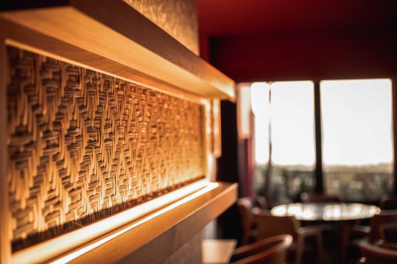 Close-up of a backlit woven rattan wall panel framed in warm oak beside sunlit windows at Al Hayat