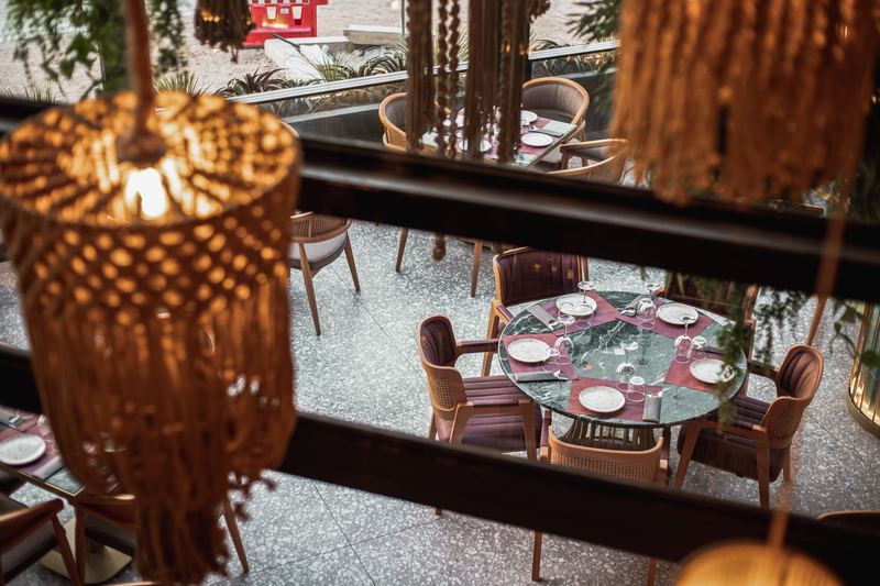 Overhead view through hanging macramé pendant lamps onto green marble tables and cane armchairs on the terrazzo dining floor