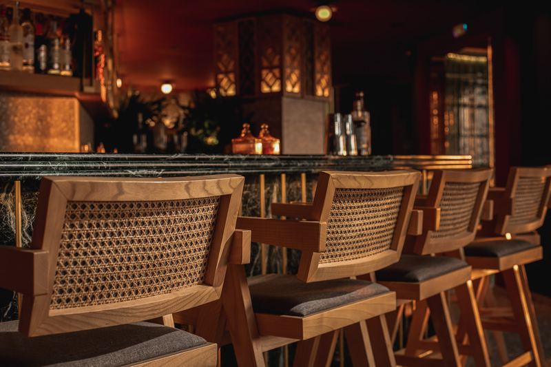 Row of cane-backed wooden bar stools lined up along Al Hayat's green marble bar under warm amber lighting
