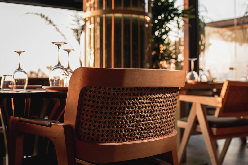 Close-up of a curved cane-back armchair beside a set table glowing in the golden sunset light streaming through the windows