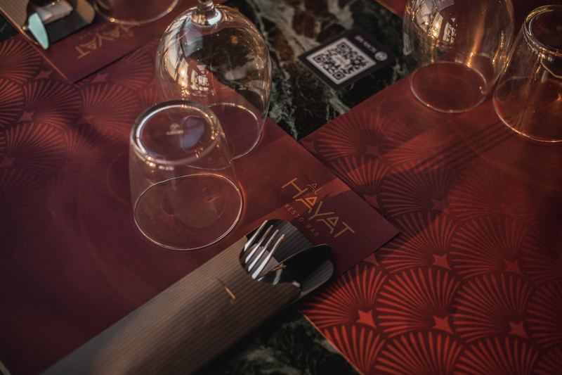 Deep red Al Hayat Resto Bar placemat with gold-foil logo, wine glasses and cutlery set on a green marble table