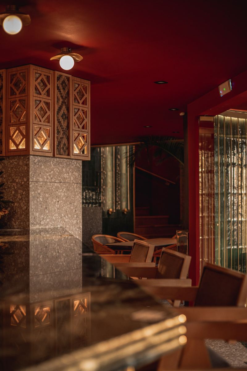 Ornate carved-wood lattice pillar glowing warm gold beside terrazzo, green tile and the staircase inside Al Hayat's crimson lounge