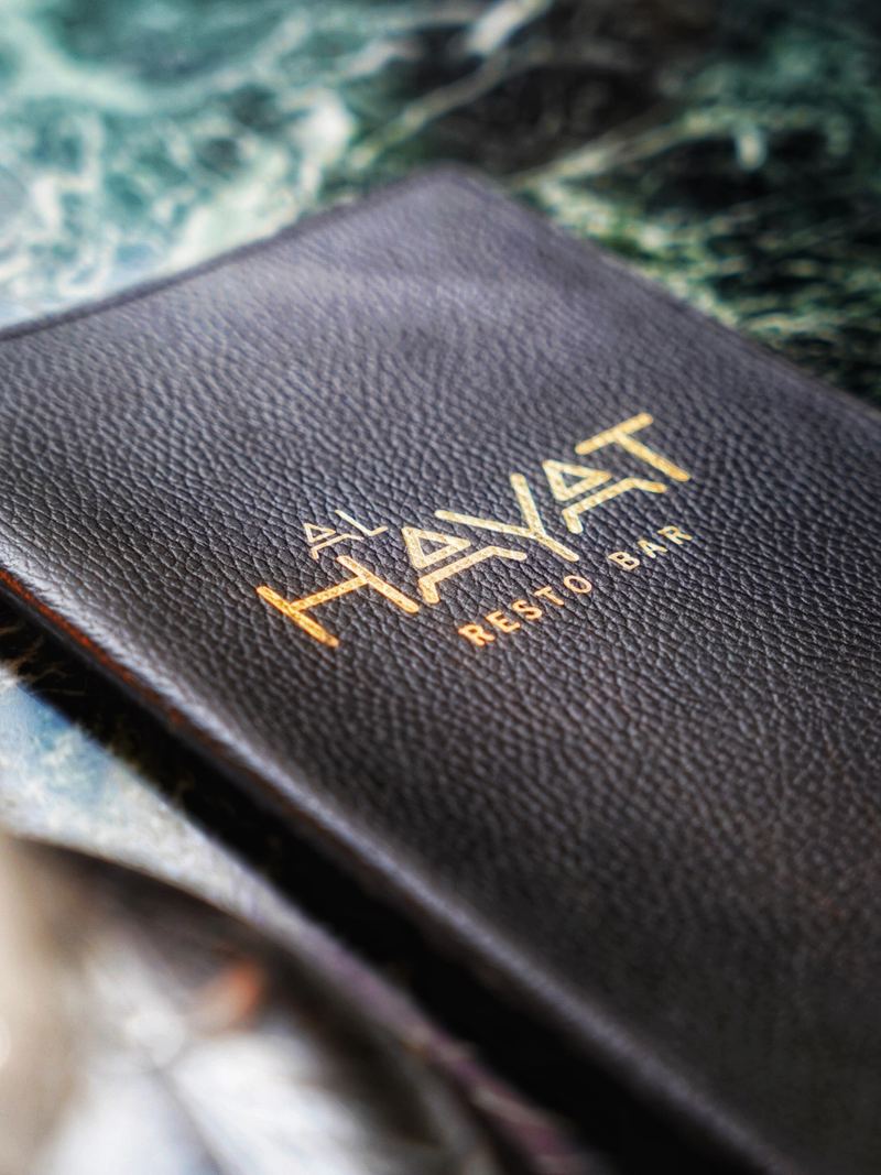 Al Hayat leather menu book with gold-foil logo resting on a green marble table