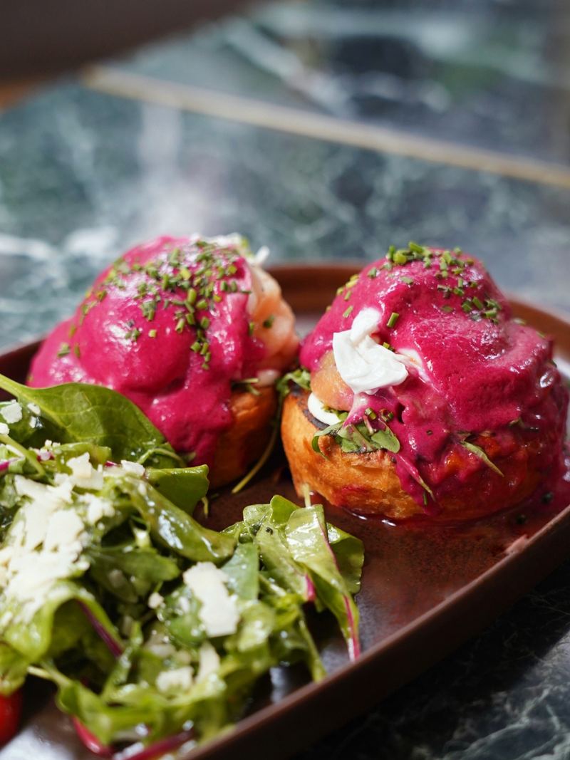 Poached eggs on brioche topped with vivid beetroot hollandaise, smoked salmon and chives, served with a fresh spinach salad