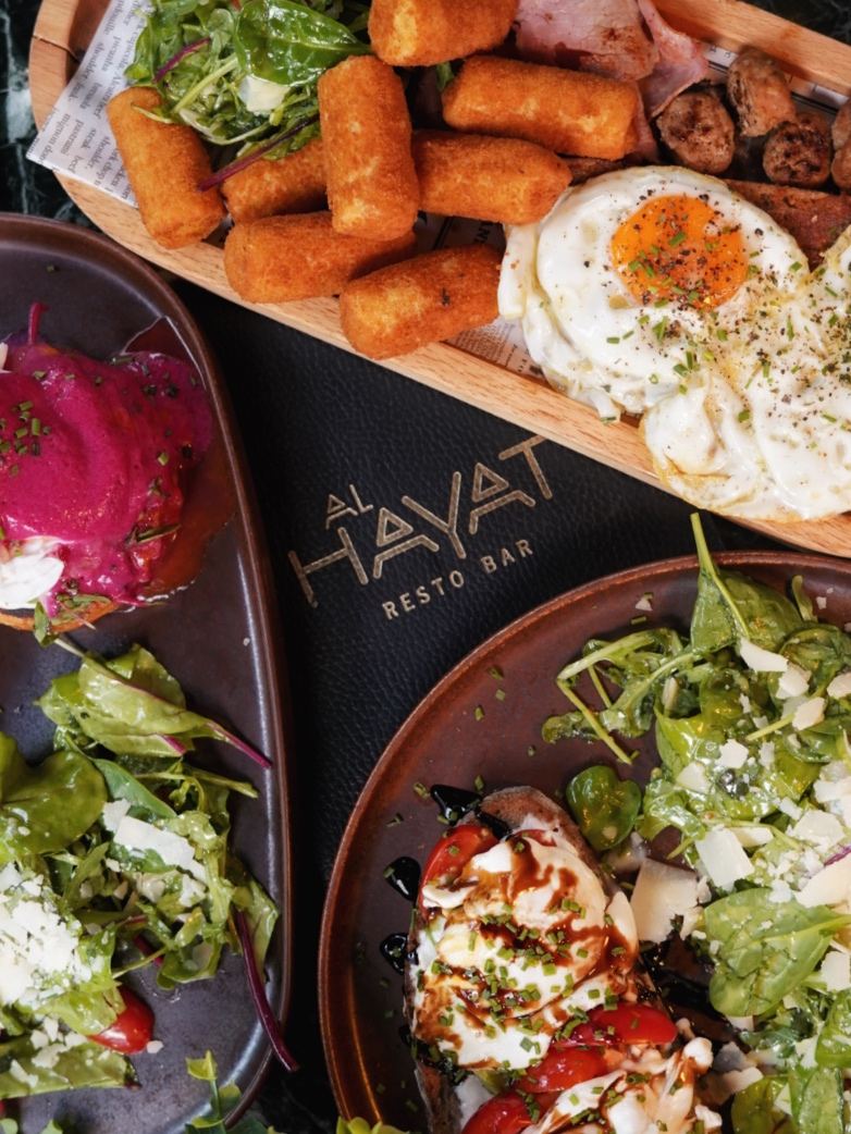 Overhead brunch spread with the Al Hayat menu, croquettes, bacon, fried eggs, beetroot benedict and burrata toast on green marble