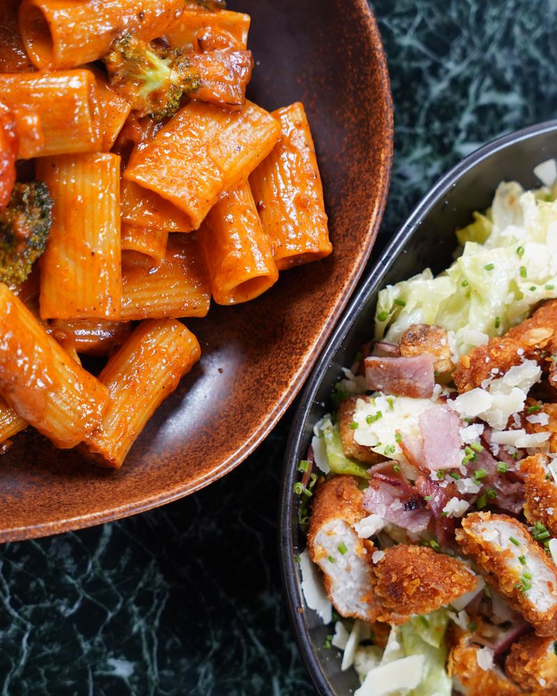 Rigatoni in spicy tomato sauce with broccoli next to a crispy chicken caesar salad with bacon and parmesan