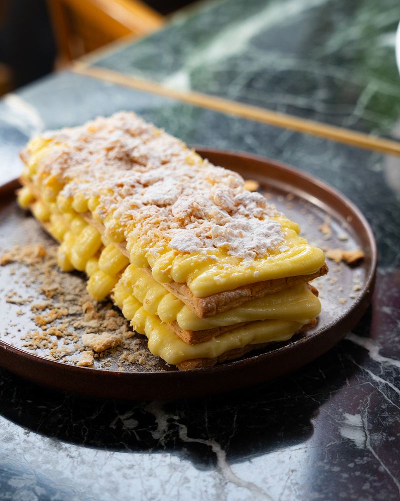 Classic mille-feuille with layers of flaky puff pastry, vanilla custard and dusted icing sugar on a dark ceramic plate