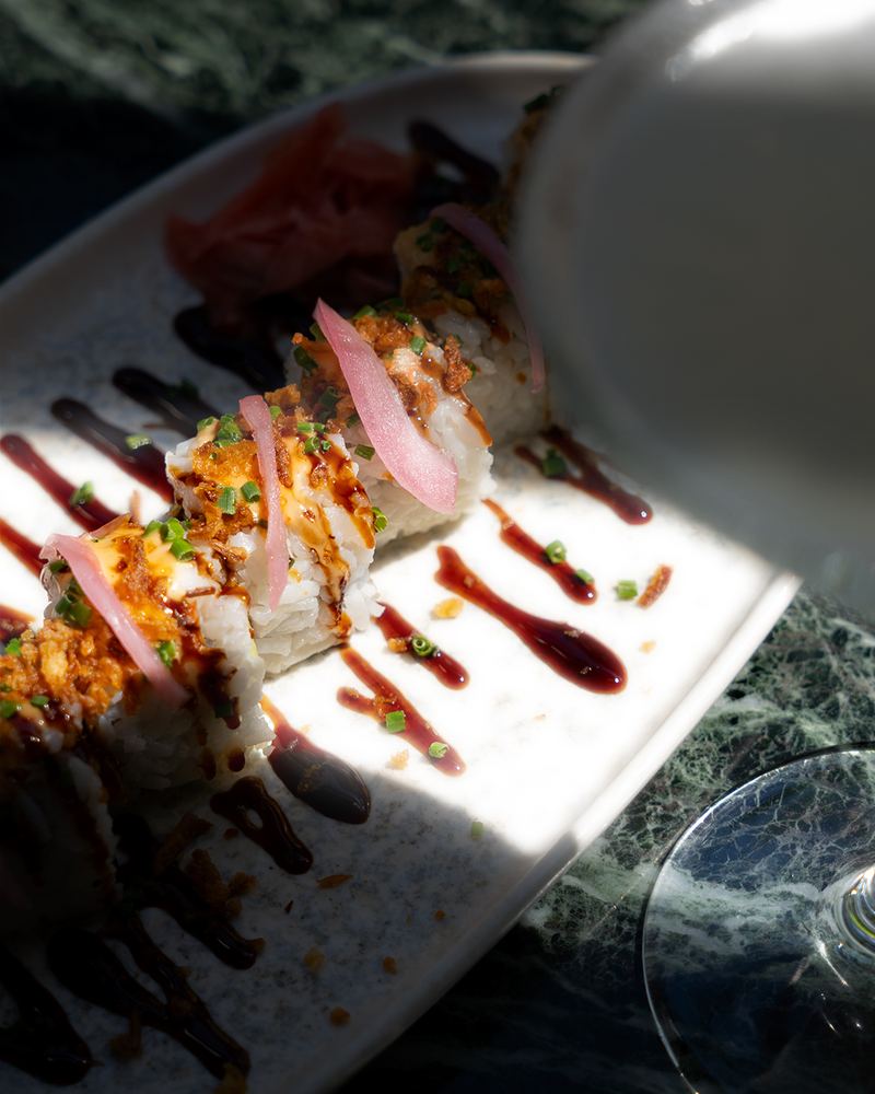 Sunlit sushi roll topped with spicy mayo, teriyaki drizzle, pickled onion and crispy garlic on a white plate over green marble