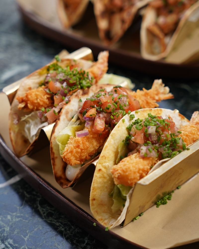 Crispy panko shrimp soft tacos with lettuce, pico de gallo and chives served on brass taco holders